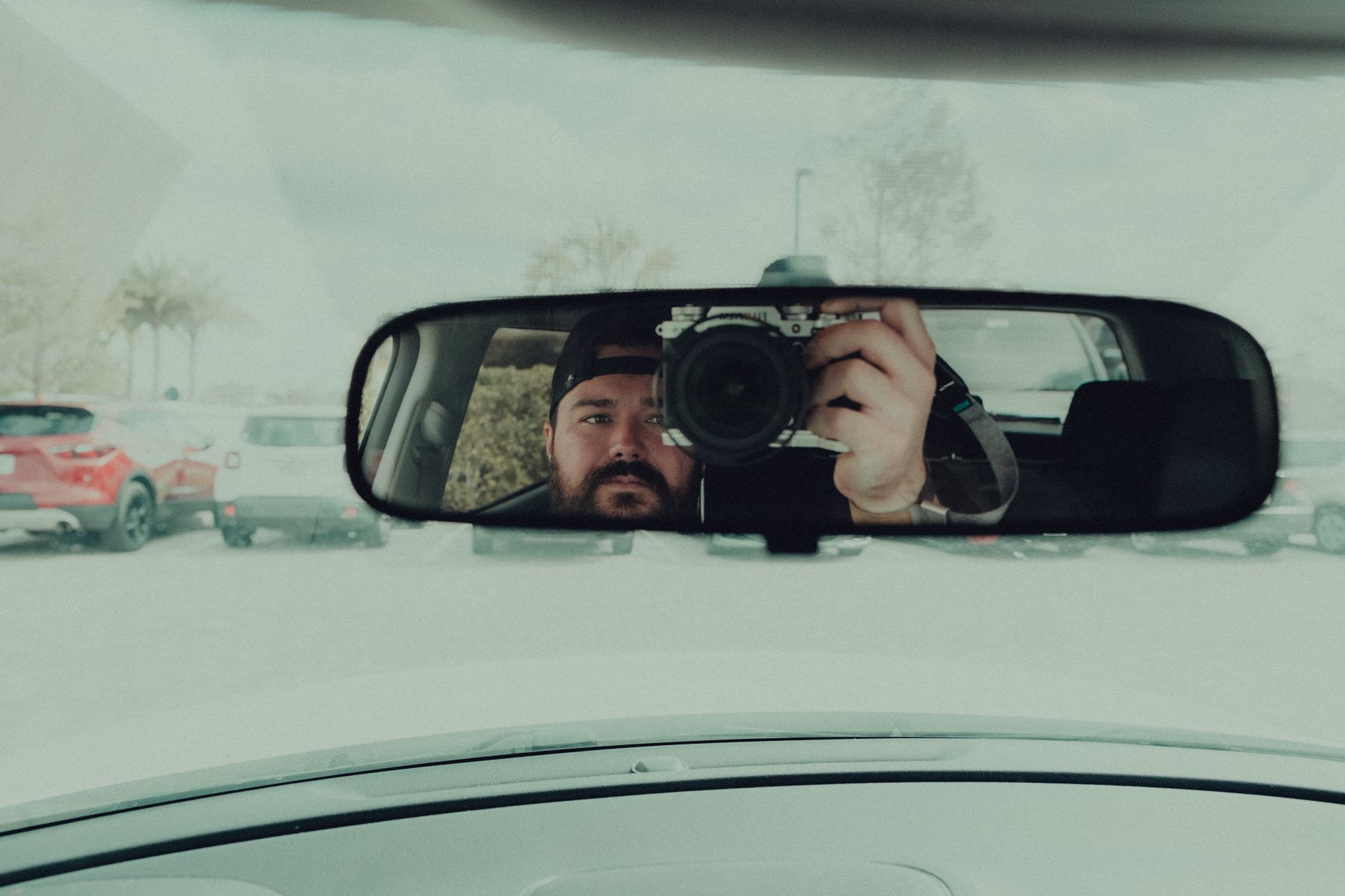 A man captures his reflection in a car's rearview mirror using a camera in a parking lot.
