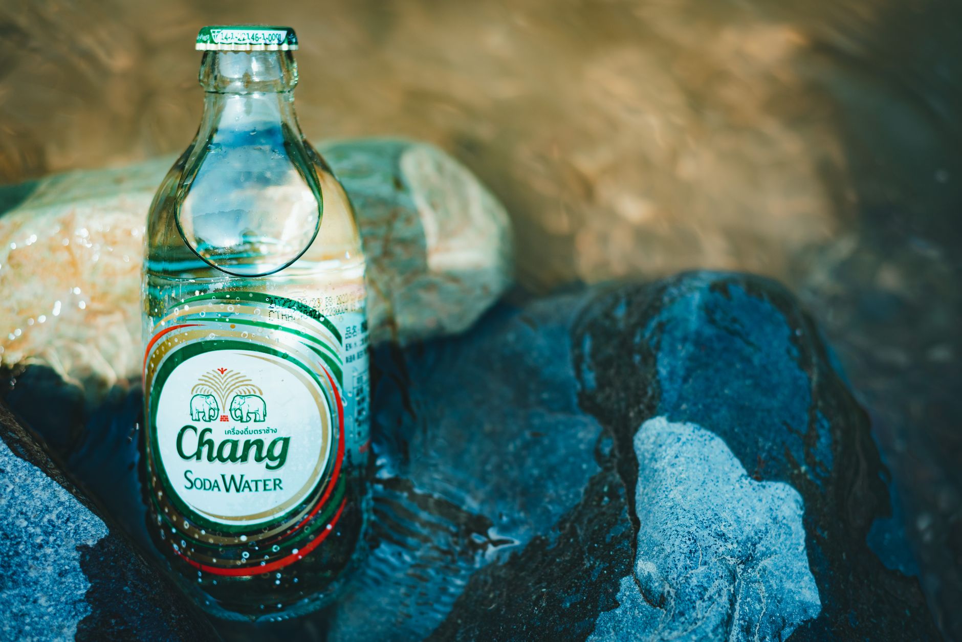 A close-up of a Chang Soda Water bottle nestled among rocks and flowing water.