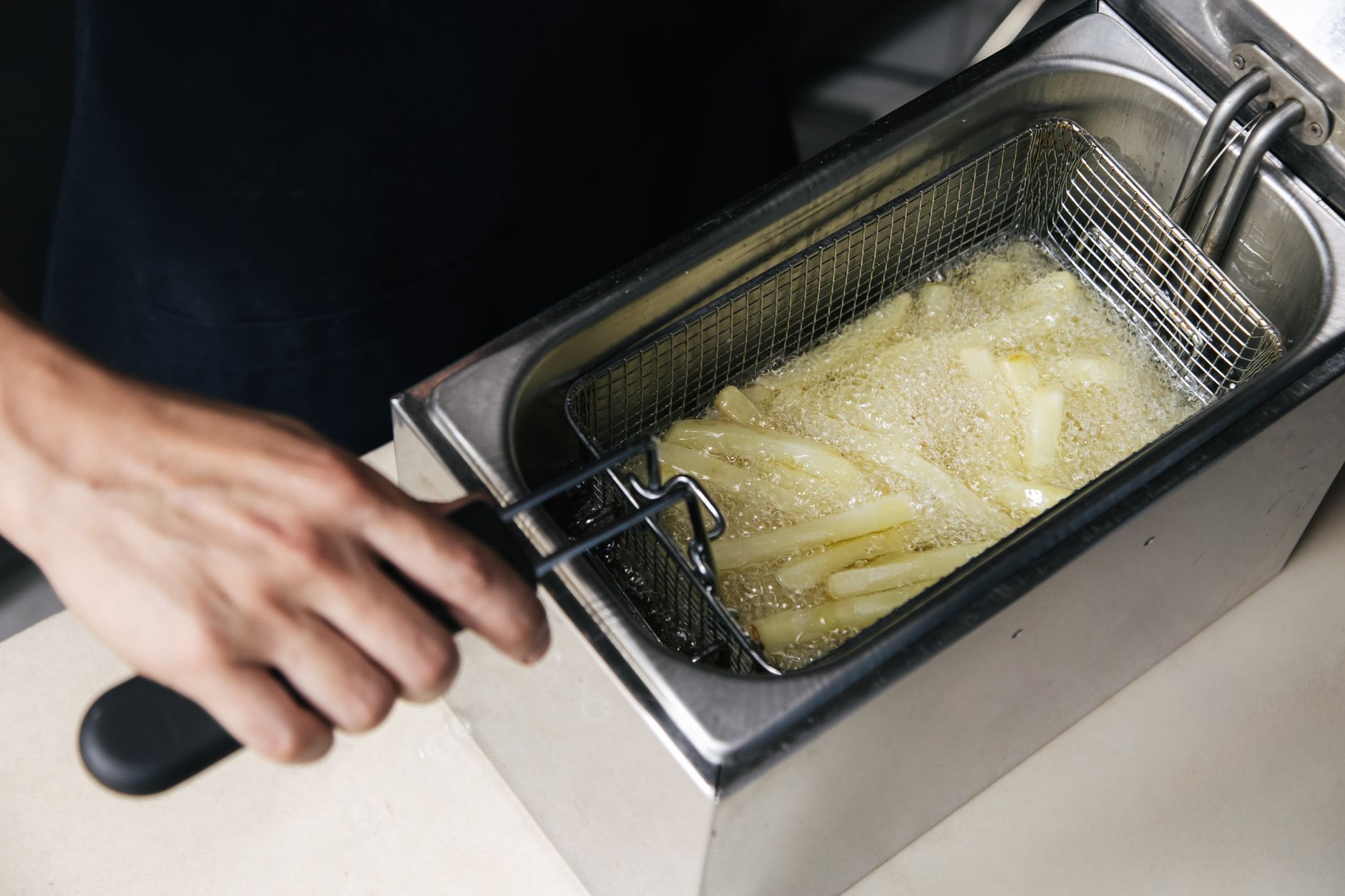 A hand operates a deep fryer cooking fresh french fries in oil.