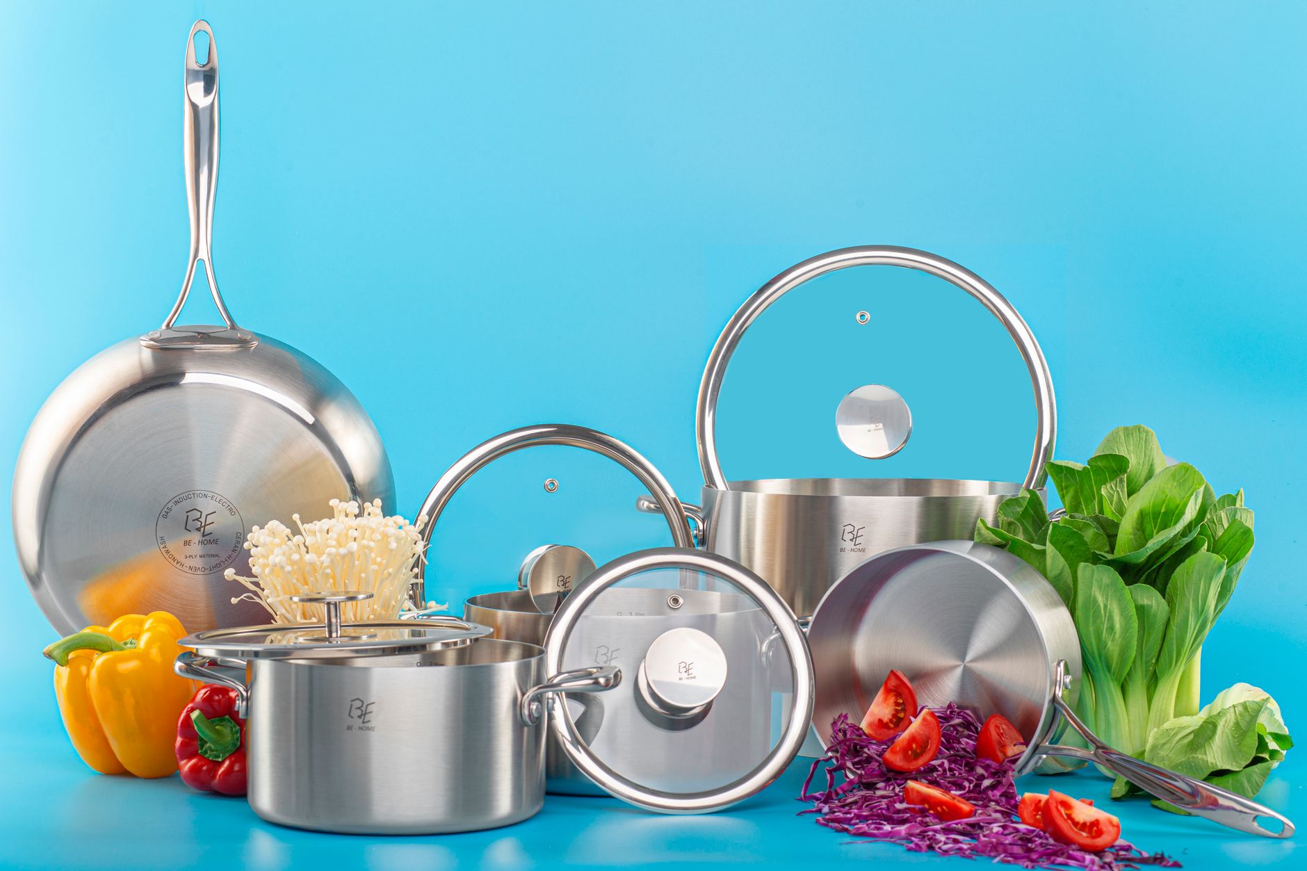 High-quality stainless steel pots and pans with fresh vegetables on a bright blue background.