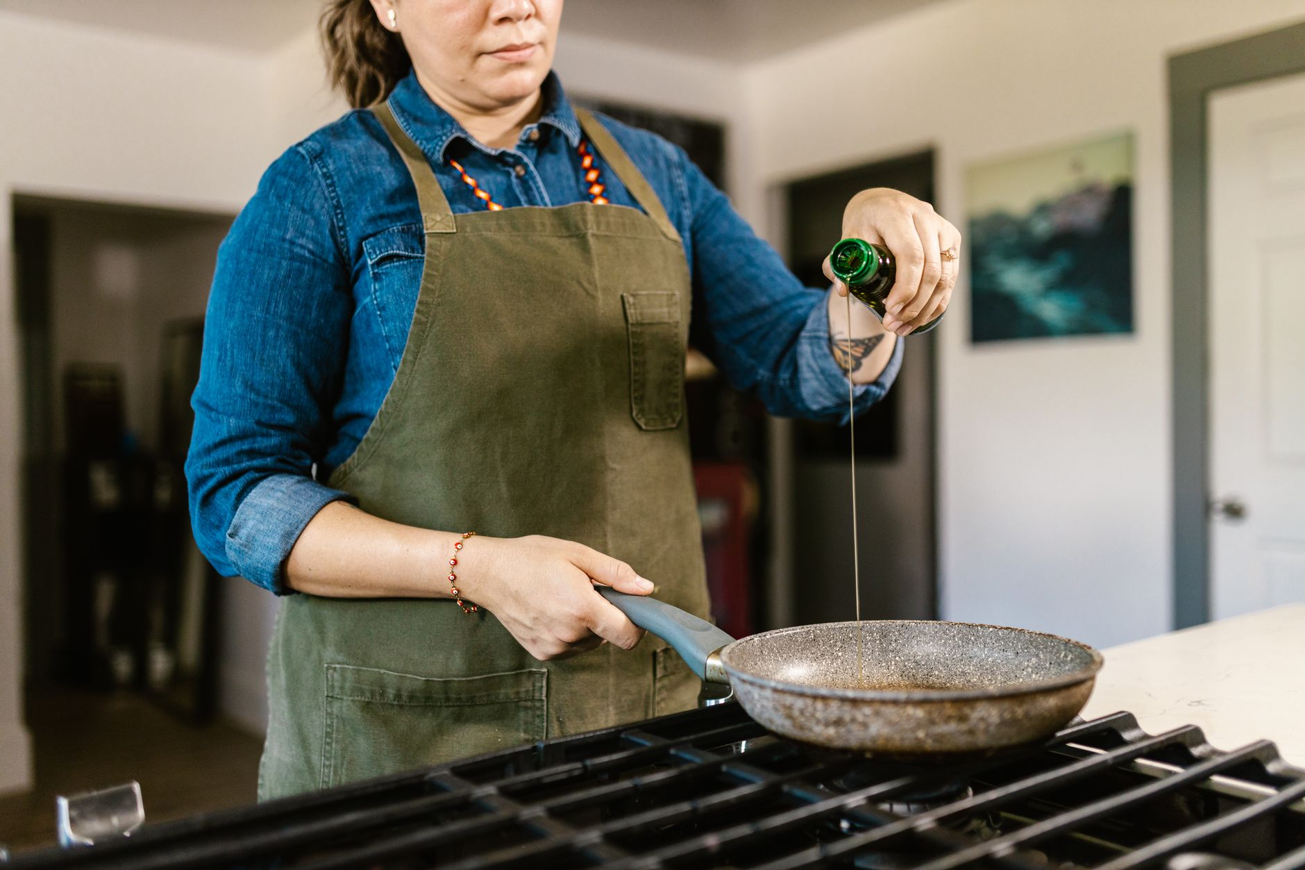 A home chef in an apron pours olive oil into a frying pan on a stove.