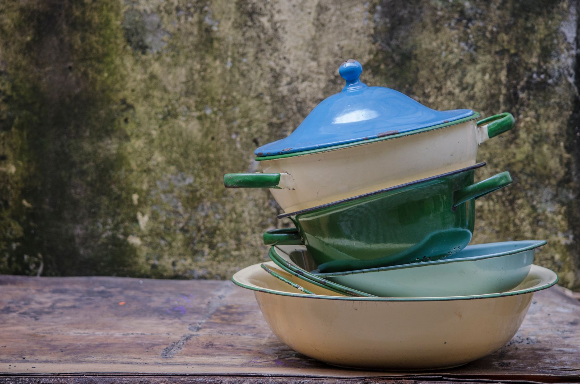 A rustic stack of colorful vintage enamel pots and pans outdoors against a textured wall.