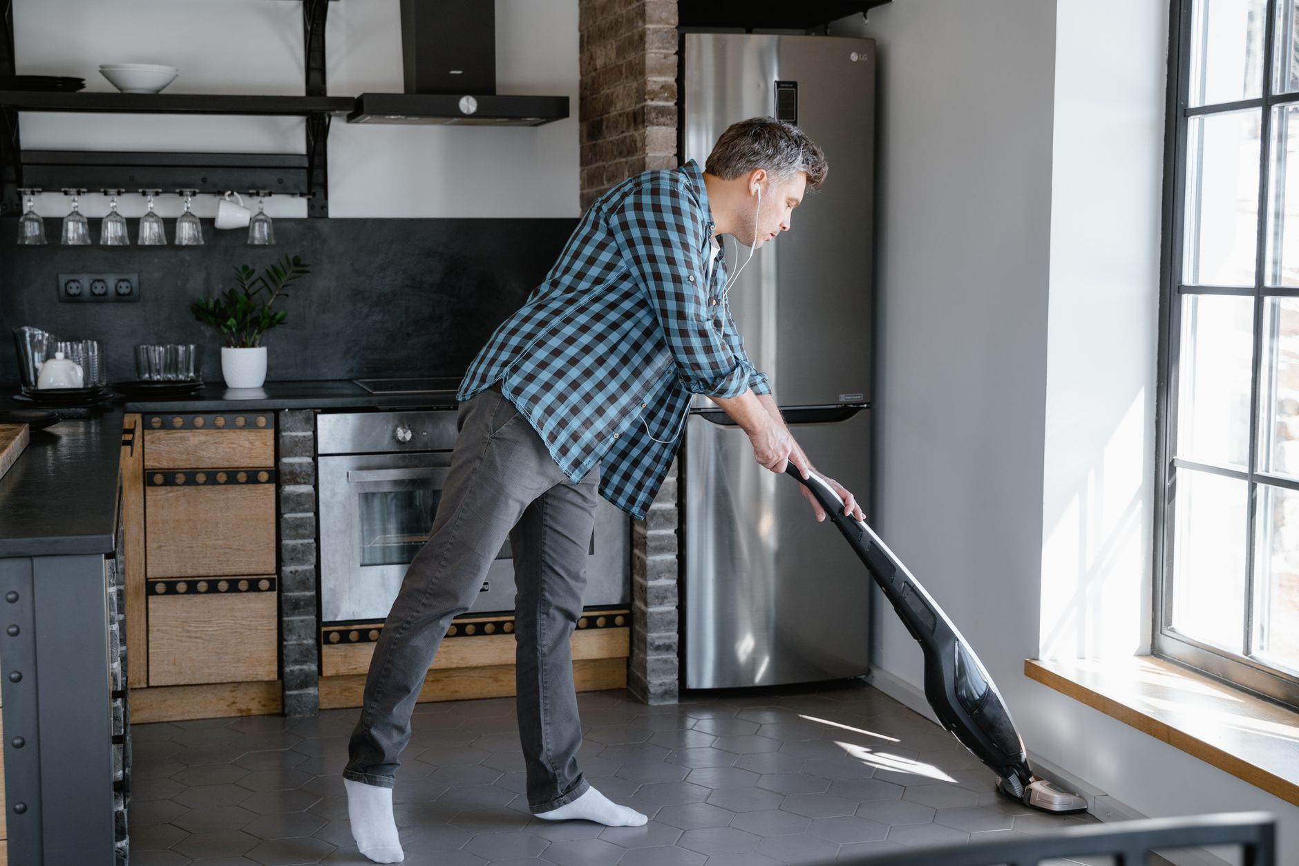 Adult man wearing earphones while vacuuming the kitchen floor at home, adding a modern lifestyle twist to household chores.