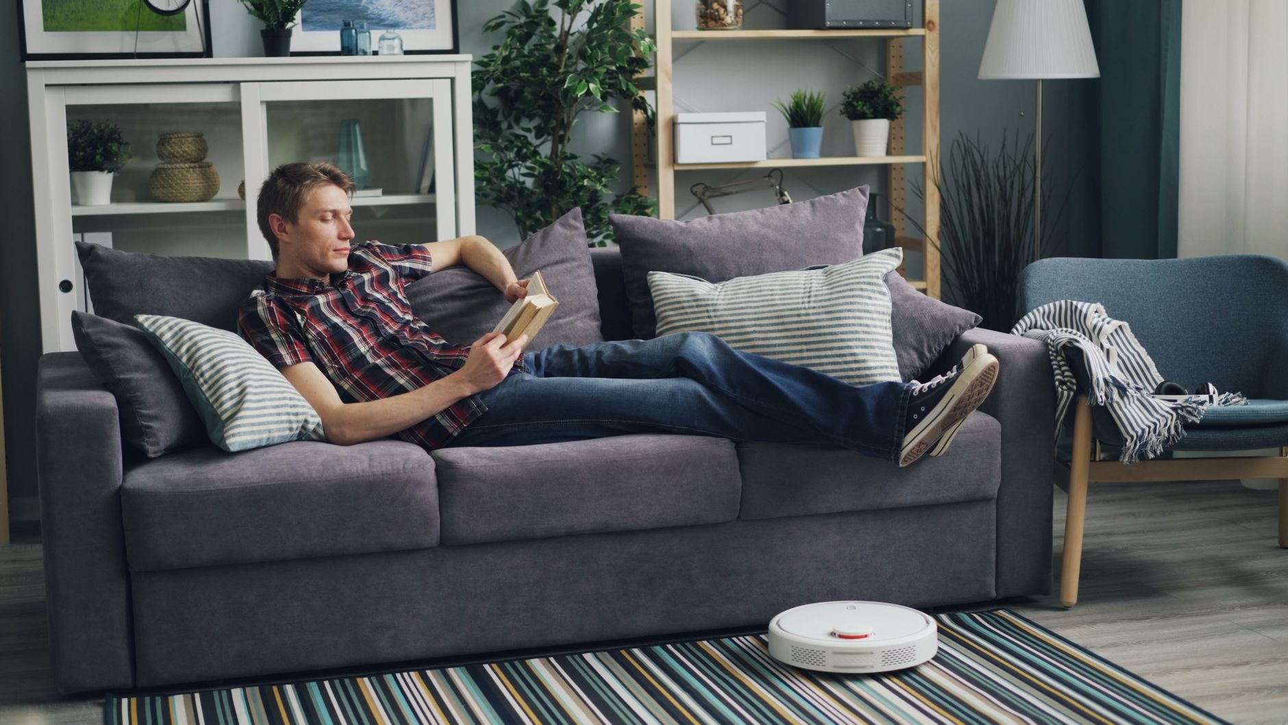 A man reading a book on a sofa, while a robotic vacuum cleans the living room floor.