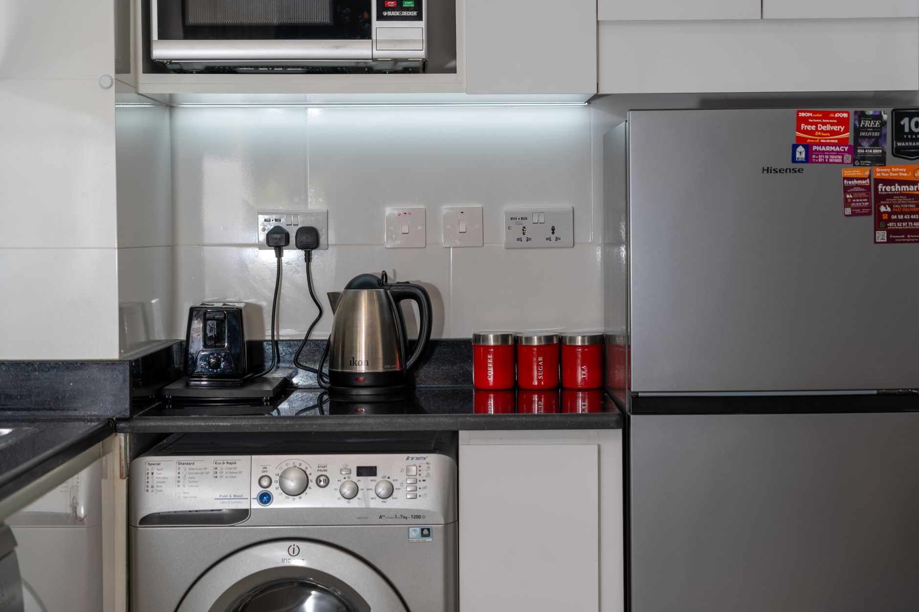 Stylish modern kitchen with stainless steel appliances and red accents, highlighting a washing machine, fridge, and countertop electronics.